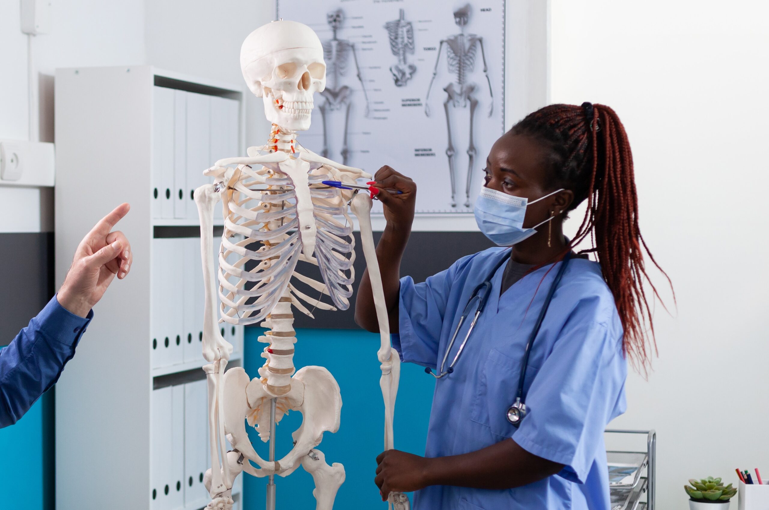African american woman nurse with medical protective face mask against covid19 holding human skeleton model explaining spine pain to senior man patient in hospital office. Osteoporosis concept