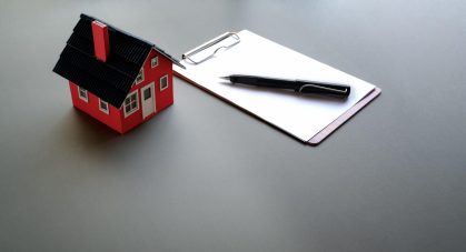 A photo of a small, model house which is red, sitting next to a clipboard with paper and a pen on it.